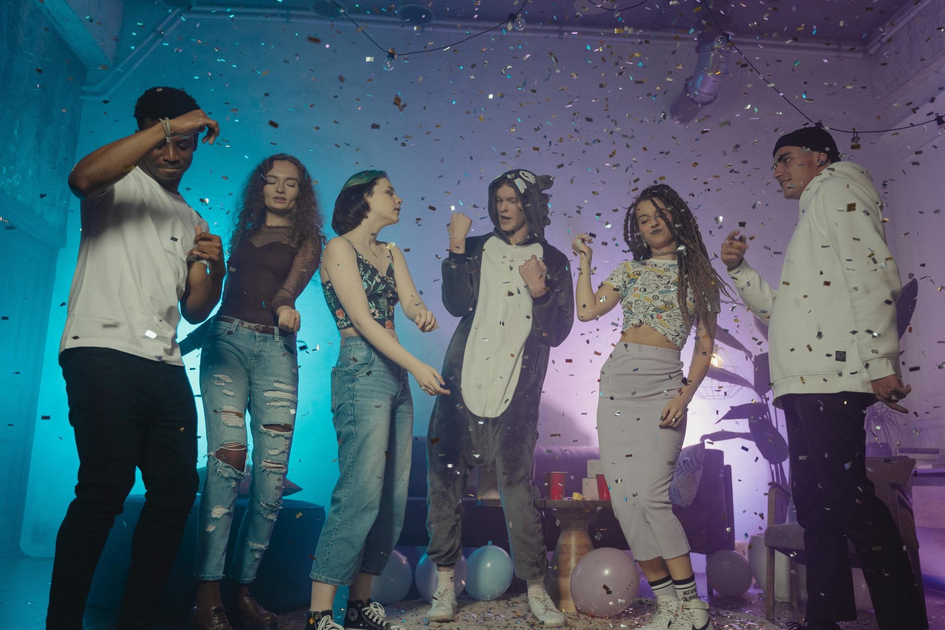 A group of diverse young adults dancing and having fun at an indoor party with confetti and balloons.